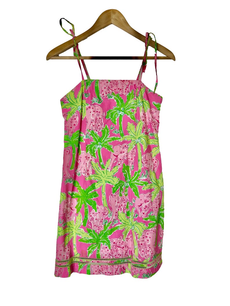 May include: Pink sundress with a tropical pattern of pink elephants and green palm trees. The dress has tie straps and a scalloped hem. The dress is hanging on a wooden hanger.