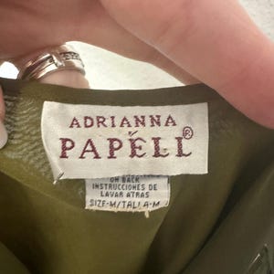 K&ouml;nnte beinhalten: Nahaufnahme eines Kleidungsst&uuml;cketiketts mit dem Markennamen "ADRIANNA PAPELL" in bordeauxroten Buchstaben. Das Etikett enth&auml;lt auch Waschanweisungen auf Spanisch und die Gr&ouml;&szlig;e "M". Der Stoff ist olivgr&uuml;n.