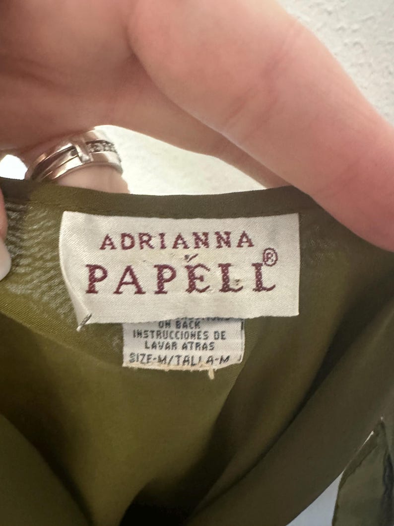 K&ouml;nnte beinhalten: Nahaufnahme eines Kleidungsst&uuml;cketiketts mit dem Markennamen "ADRIANNA PAPELL" in bordeauxroten Buchstaben. Das Etikett enth&auml;lt auch Waschanweisungen auf Spanisch und die Gr&ouml;&szlig;e "M". Der Stoff ist olivgr&uuml;n.