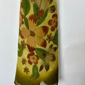 K&ouml;nnte beinhalten: Ein langes Kleid mit floralem Muster. Das Kleid zeigt gro&szlig;e, stilisierte Blumen in Braun-, Orange- und Gelbt&ouml;nen mit gr&uuml;nen Bl&auml;ttern auf einem hellgelben Hintergrund. Der Stoff wirkt transparent und leicht.