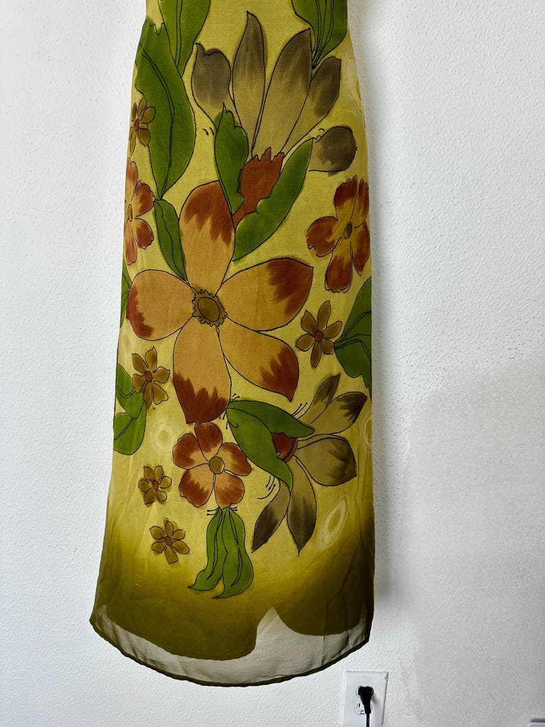 K&ouml;nnte beinhalten: Ein langes Kleid mit floralem Muster. Das Kleid zeigt gro&szlig;e, stilisierte Blumen in Braun-, Orange- und Gelbt&ouml;nen mit gr&uuml;nen Bl&auml;ttern auf einem hellgelben Hintergrund. Der Stoff wirkt transparent und leicht.