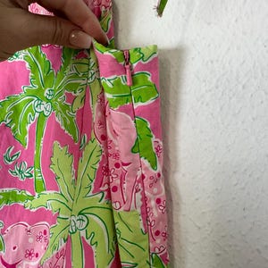Op de afbeelding: Felroze stof met een groen tropisch patroon van palmbomen en olifanten. De stof wordt gebruikt voor een kledingstuk, met een rits en bandjes zichtbaar. Het kleurenschema is helder en zomers.