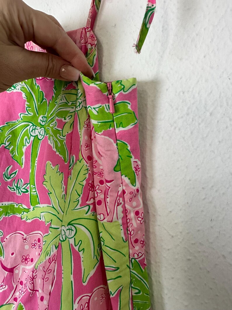 Op de afbeelding: Felroze stof met een groen tropisch patroon van palmbomen en olifanten. De stof wordt gebruikt voor een kledingstuk, met een rits en bandjes zichtbaar. Het kleurenschema is helder en zomers.