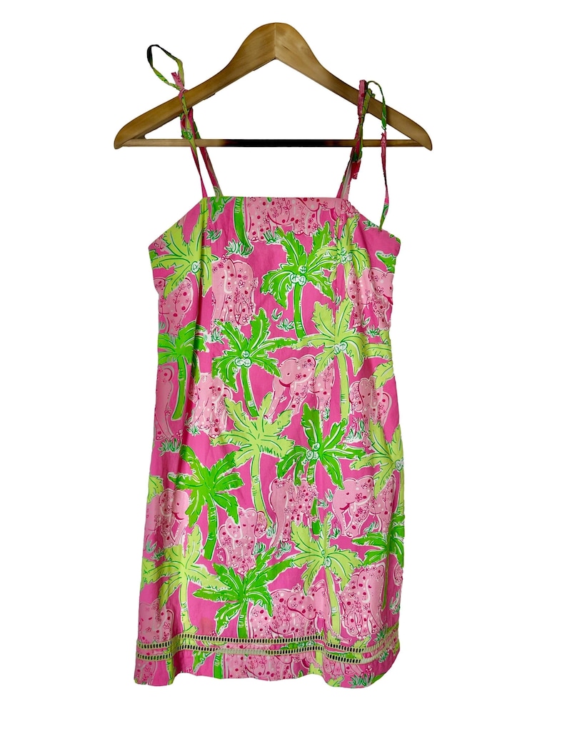 Op de afbeelding: Roze zomerjurk met een tropische print met olifanten en groene palmbomen. De jurk heeft strikbandjes en een geschulpte zoom. De jurk hangt aan een houten kleerhanger.