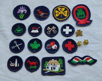 Puede incluir: Una colección de 15 parches bordados, la mayoría azules con costuras blancas, que presentan varios diseños, incluyendo una hoja de arce, una casa, una cruz, una llave, una máquina de coser, una taza, un pájaro, un trébol, un logotipo de guía y un par de tijeras.