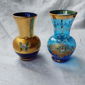 May include: Two decorative glass vases. One is gold with blue accents and floral designs. The other is light blue with gold trim and floral patterns. Both vases have a bulbous base and a flared top.