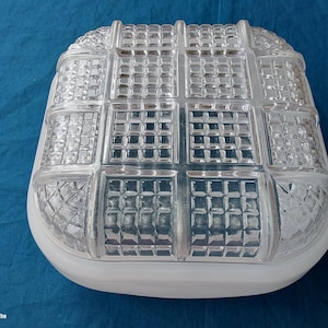 May include: A clear, square-shaped glass light fitting with a grid pattern. The fitting has a white, rounded base and a textured, geometric design on the glass. The light fitting is likely for indoor or outdoor use.
