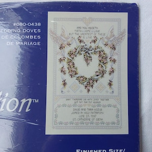 May include: A white cross-stitch pattern featuring two doves and a heart-shaped wreath. The pattern includes the text "And now abideth faith, hope, love, but the greatest of these is love." and "What therefore God hath joined together let not man put asunder."  The pattern also includes the names "David and Tara Kozub" and the date "June 27, 1997".