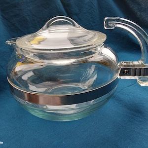 Könnte beinhalten: Eine klare Glasteekanne mit einem silbernen Band um die Mitte und einem Glasdeckel. Die Teekanne hat einen Glasgriff.