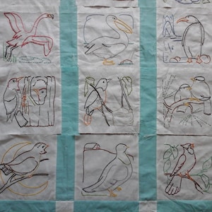 May include: A handmade quilt with embroidered bird designs. Each square features a different bird, such as a flamingo and pelican, outlined in black thread with red, orange, and yellow accents. The quilt has light blue vertical stripes.