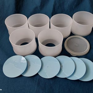 Może przedstawiać: Zestaw ośmiu białych plastikowych pojemników o matowym wyglądzie. Siedem cylindrycznych kubków ułożonych wokół pojedynczej pokrywki i sześciu jasnoniebieskich okrągłych pokrywek. Przedmioty znajdują się na ciemnoniebieskiej powierzchni.