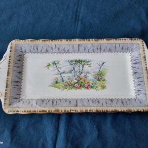May include: A rectangular ceramic serving tray with a white centre featuring a colourful landscape illustration of birch trees and wildflowers. The border has a grey birch bark pattern with gold trim on the scalloped handles and edges.