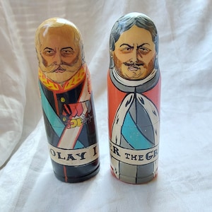 May include: Two wooden nesting dolls, or matryoshka dolls, painted with portraits of historical figures. The doll on the left is painted with a portrait of a man with a moustache and beard, and the text "OLAY I" is written on the bottom. The doll on the right is painted with a portrait of a man with a moustache and the text "ER THE GR" is written on the bottom.