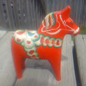 May include: A bright red wooden Dala horse, a traditional Swedish folk art item. It features painted floral patterns in white, green, and blue. The horse has a detailed mane and tail. A charming decorative piece.