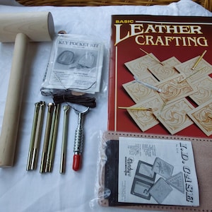 May include: A leather crafting kit featuring a wooden mallet, metal tools, a key pocket kit, and a book titled "Basic Leather Crafting". The book is red with gold lettering and illustrations of leather tooling.