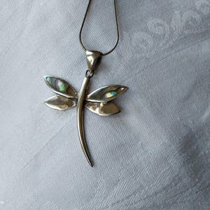 May include: Silver dragonfly pendant necklace. The dragonfly pendant has iridescent abalone shell accents on the wings. The necklace is displayed on a white, textured surface.