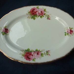 May include: An oval, white porcelain serving platter with a scalloped edge and gold trim. The platter is decorated with clusters of pink roses and green leaves. A classic piece for serving food.