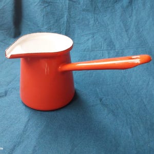 Peut inclure: Petite cafetière en émail rouge avec une longue poignée orange. L'intérieur et le bord du pot sont blancs. La cafetière est conçue pour verser et a un aspect classique et vintage.