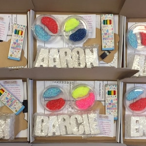 Puede incluir: Seis cajas con kits de manualidades para niños. Cada caja contiene un conjunto de cuentas de plástico de colores, un pequeño recipiente de purpurina, un conjunto de mini lápices y un conjunto de letras de espuma blanca que deletrean un nombre. Los nombres son: 'Aaron', 'Lucas', 'Indiah', 'Darcie', 'Emma' y 'Ceasyn'.