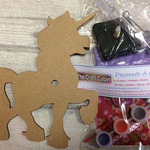 Può includere: Un kit per orologio di legno a forma di unicorno con un meccanismo di orologio, colori, glitter, gemme, colla e un pennello. Il kit include istruzioni per decorare l'orologio. "Decorate A Clock Kit" e "The Craft Cabin" sono stampati sulla confezione.