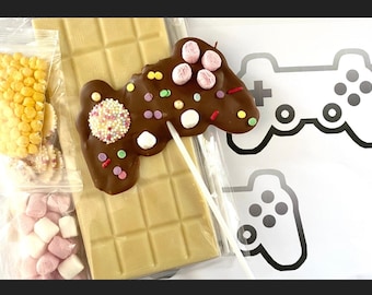 May include: A white chocolate bar with a chocolate game controller on a stick. The game controller is decorated with sprinkles, marshmallows, and candy. There are bags of sprinkles and marshmallows next to the chocolate bar.