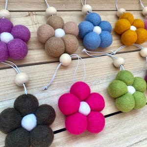 May include: Ten felt flower ornaments in various colors, including pink, purple, brown, blue, green, yellow, and burgundy. Each flower has a white center and is attached to a wooden bead and a white string.