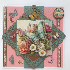 Op de afbeelding: Decoratieve wenskaart met een roze achtergrond en een centraal ontwerp met een grote vlinder met blauwe en gouden accenten, omringd door bloemen. De kaart bevat de tekst "Flutterbye Wishes".