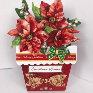 以下が含まれることがあります： 赤い鉢にポインセチアの花がアレンジされた赤と緑のクリスマスカード。鉢は白い波状の縁取りと「メリークリスマス」と書かれた金色のリボンが付いています。鉢には金色のリボンと「クリスマスの願い」の文字も書かれています。