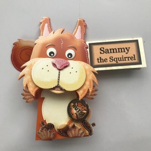 Puede incluir: Un títere de ardilla marrón y naranja con un cartel que dice "Sammy la ardilla". La ardilla sostiene una moneda de oro que dice "Go Nuts!"