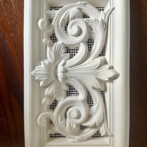 Op de afbeelding: Een rechthoekige, crèmekleurige decoratieve ventilatieafdekking met een bloemen- en krulontwerp. Het centrale ontwerp heeft een grote bloem en wervelende bladeren, tegen een zwarte gaasachtergrond. Het frame is glad en wit geschilderd.