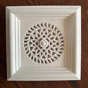 May include: A square, off-white vent cover featuring a central circular cutout with an intricate, geometric star-like pattern. The frame has a beveled edge, and the circular design includes a small, raised circular element at its centre.