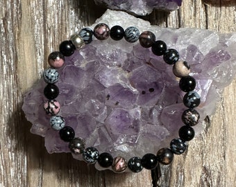 May include: A black and pink beaded bracelet with a silver charm. The bracelet is on a bed of amethyst crystals.