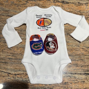 May include: White baby onesie with long sleeves and the text "Daddy loves F, Mommy loves F, But They Both Love ME." Includes baby shoes, one blue with a Florida Gators logo and one maroon with a Seminoles logo.