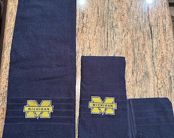 May include: A set of three navy blue towels with a textured surface. Each towel features an embroidered yellow and blue block M logo with the word MICHIGAN printed in black text across the center of the letter, positioned near the bottom edge.
