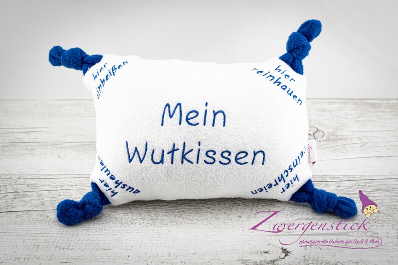 Puede incluir: Almohada blanca con esquinas azules anudadas y el texto "Mein Wutkissen" bordado en la parte delantera. La almohada tiene el texto "hier inbei&szlig;en", "reinhauen", "hier ausheulan" y "hier reinschreien" bordado en los lados.
