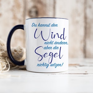 Può includere: Tazza in ceramica bianca con bordo e manico blu. La tazza presenta un design blu e bianco con il testo "Du kannst den Wind nicht ändern, aber die Segel richtig setzen!"