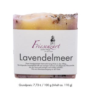 Könnte beinhalten: Ein Stück handgemachte Seife mit einem weißen Etikett mit dem Text "Friesenzart Lavendelmeer". Die Seife hat eine hellbraune und violette Farbe mit sichtbaren Pflanzenbestandteilen. Das Etikett enthält auch eine Leuchtturm-Illustration.