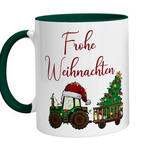 Può includere: Tazza in ceramica bianca con bordo verde. La tazza presenta un'illustrazione rossa e bianca di un trattore che tira un albero di Natale. Il testo "Frohe Weihnachten" è stampato in rosso sulla tazza.