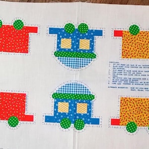 May include: A white fabric panel featuring colorful train car designs. The train cars are in various colors, including red, blue, yellow, and green, with polka dot and checkered patterns. The panel has a vintage, handmade aesthetic.