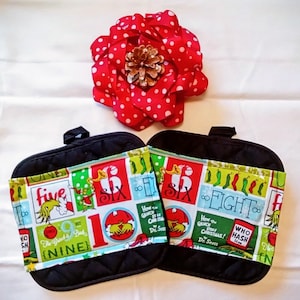 May include: Two black quilted oven mitts with colorful Dr. Seuss-themed fabric accents featuring numbers and characters. A large red polka-dot fabric flower with a pinecone center sits above the mitts.