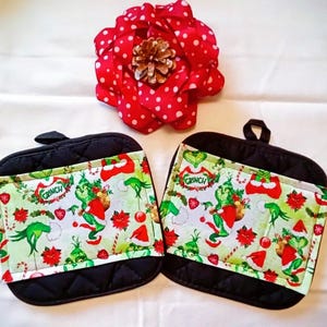 May include: Two black quilted pot holders with a Grinch-themed design. The design features the Grinch, Christmas decorations, and the word "Grinch". A red polka-dot flower with a pinecone center is in the background.