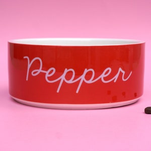 May include: Red ceramic pet bowl with the name "Pepper" written in white cursive lettering.  A few brown dog food kibbles are scattered on a pink surface.