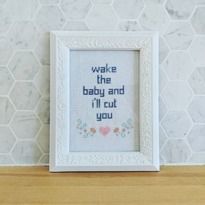 Könnte beinhalten: Weiß gerahmter Kreuzstich mit einem Blumendesign und dem Text "wake the baby and i'll cut you" in blauer Farbe.