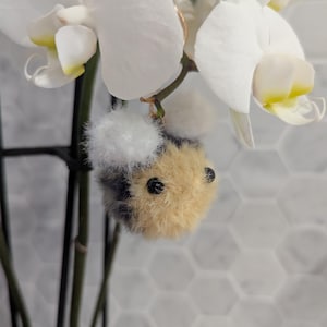 May include: A fuzzy, black, white, and brown bee keychain with black eyes. The keychain is hanging from a white orchid plant.