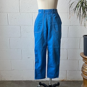 May include: Bright blue high-waisted pants with a wide leg design. The pants feature large front pockets and vertical seams down the front and back of the legs. The pants are displayed on a mannequin against a white brick wall.