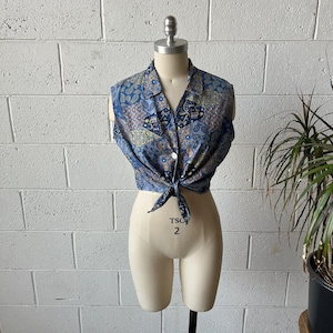May include: A sleeveless blue patterned top with a button-down collar and a tie at the waist. The fabric features a paisley and geometric design in shades of blue, yellow, and brown. The top is displayed on a mannequin.