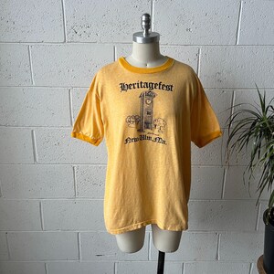 May include: Yellow Heritagefest t-shirt with a black graphic of a clock tower and the text "New Ulm, Mn." The shirt has a yellow collar and sleeves. The shirt is displayed on a mannequin.
