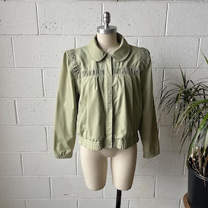 May include: A light green jacket with a rounded collar and gathered detailing across the chest and shoulders. The jacket has long sleeves, a button closure, and a gathered hem. The jacket is displayed on a mannequin against a white brick wall.