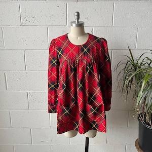 May include: A vintage red plaid dress with long sleeves and a scoop neckline. The dress features a classic plaid pattern in red, black, and yellow, with two front pockets. The dress is displayed on a mannequin against a white brick wall.