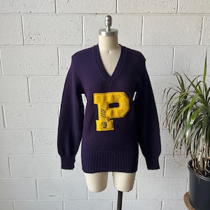 May include: A vintage purple V-neck sweater with a large yellow letter "P" on the front. The letter has a small emblem at the bottom. The sweater is displayed on a mannequin against a white brick wall.